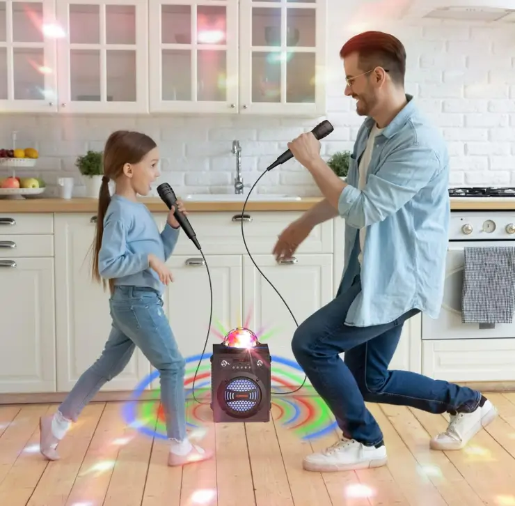 A father and daughter dancing and singing into microphones in a bright kitchen, with a light-up party speaker on the floor